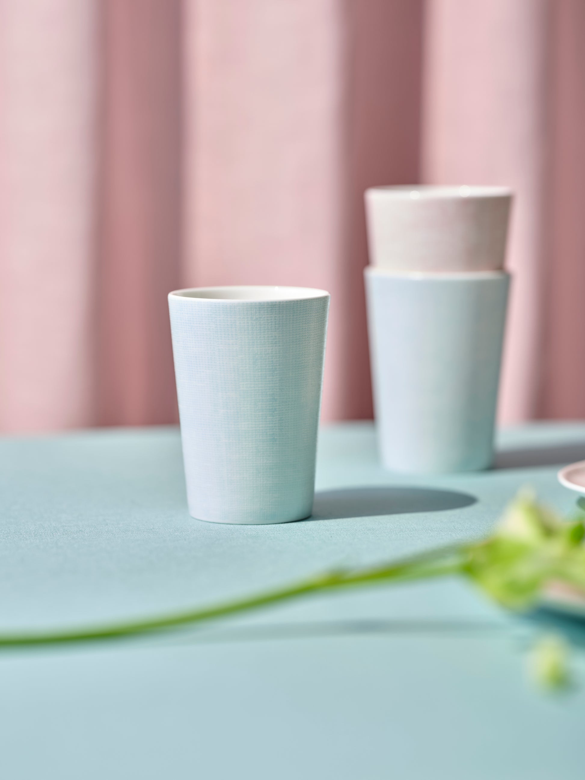 Two light blue ceramic cups on a light green surface with a pink curtain in the background.