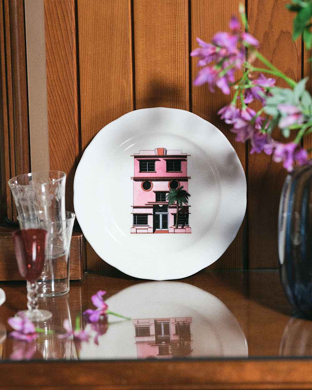 White plate with a pink building illustration on a wooden table with flowers and glasses.