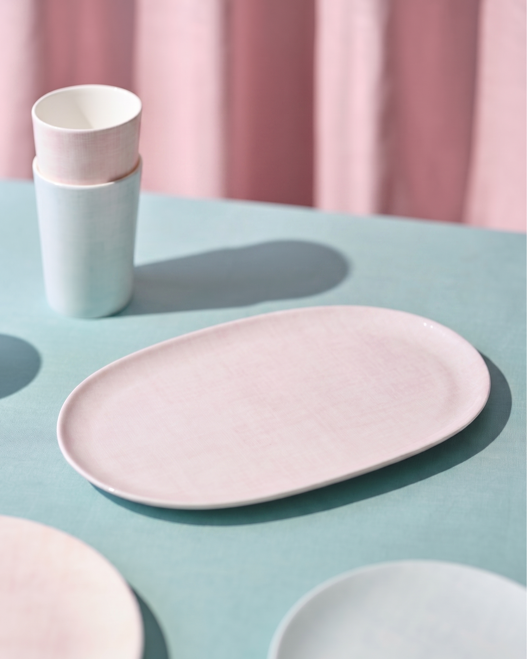 Pink ceramic plate and cups on a light blue surface with pink curtains in the background